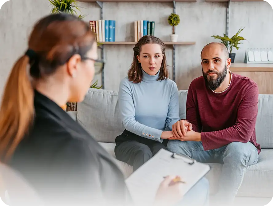 This image depicts a counselling session. In the foreground there is a counsellor taking notes. The counsellor is facing away, toward a couple, a man and a woman, sitting on a couch. The couple are holding hands and looking intently toward the counsellor.
